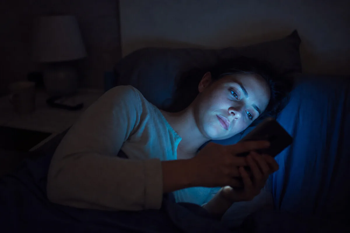 Person im Bett mit Smartphone – blaues Bildschirmlicht stört Melatoninproduktion und Tiefschlaf