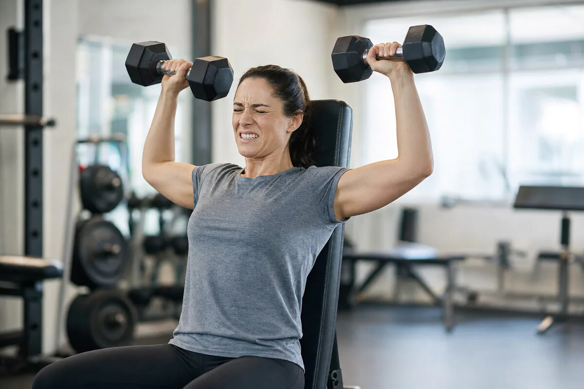 Schulterdrücken im Fitnessstudio – schmerzverzerrter Gesichtsausdruck bei Überkopfbelastung, typisch für SAS