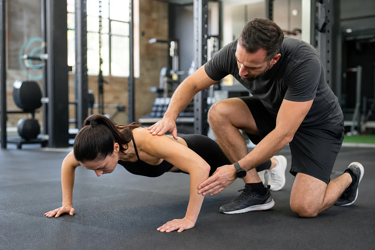 Zertifizierter Fitnesstrainer korrigiert die Liegestuetz-Technik einer Trainierenden im Studio